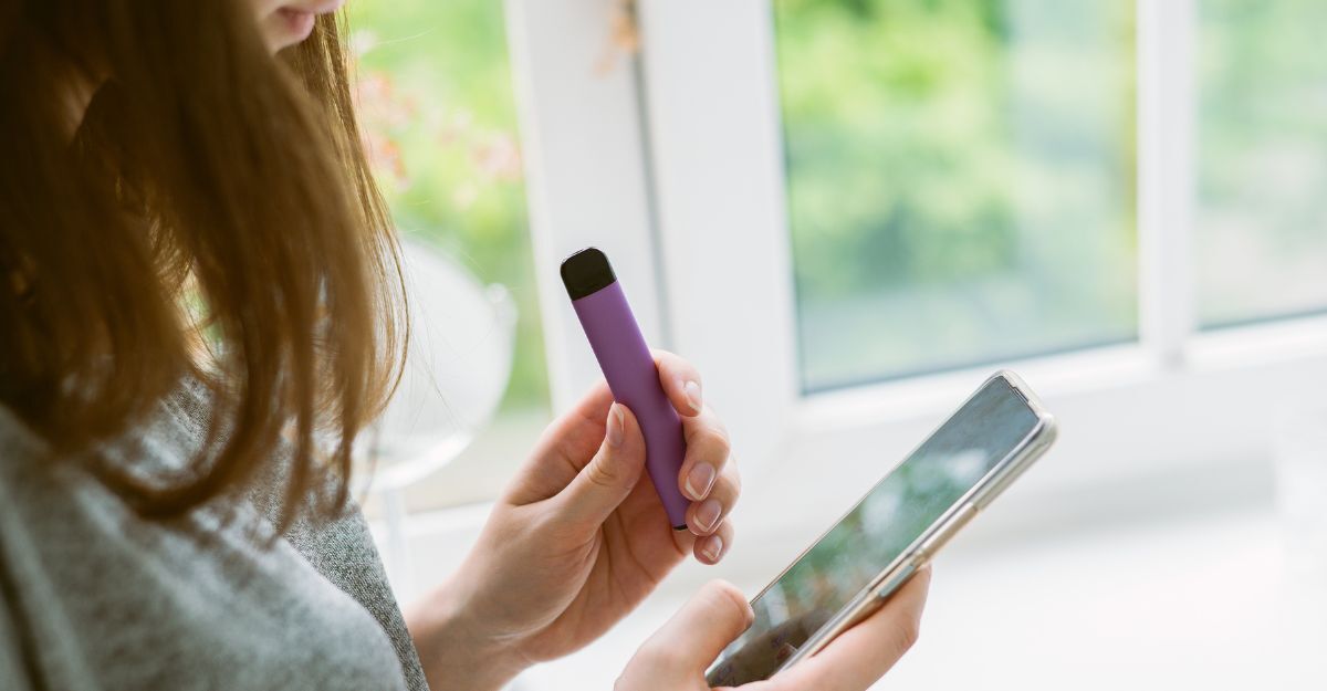 Una ragazza tiene in mano uno smartphone mentre fuma una sigaretta elettronica usa e getta viola vicino alla finestra