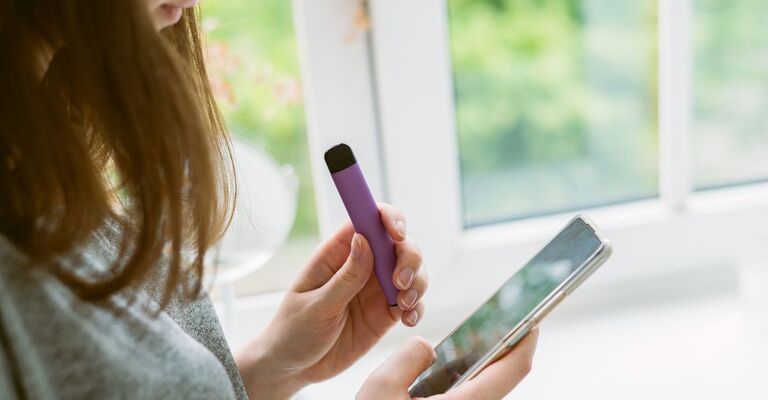 Una ragazza tiene in mano uno smartphone mentre fuma una sigaretta elettronica usa e getta viola vicino alla finestra