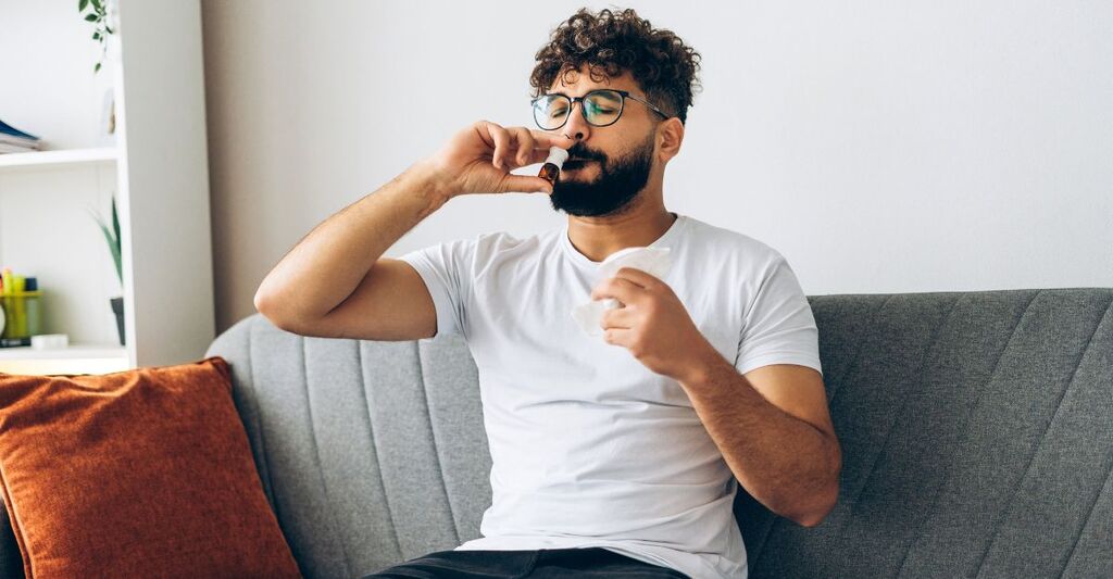 Un uomo mediorientale triste usa uno spray nasale per curare le allergie a casa, seduto sul divano