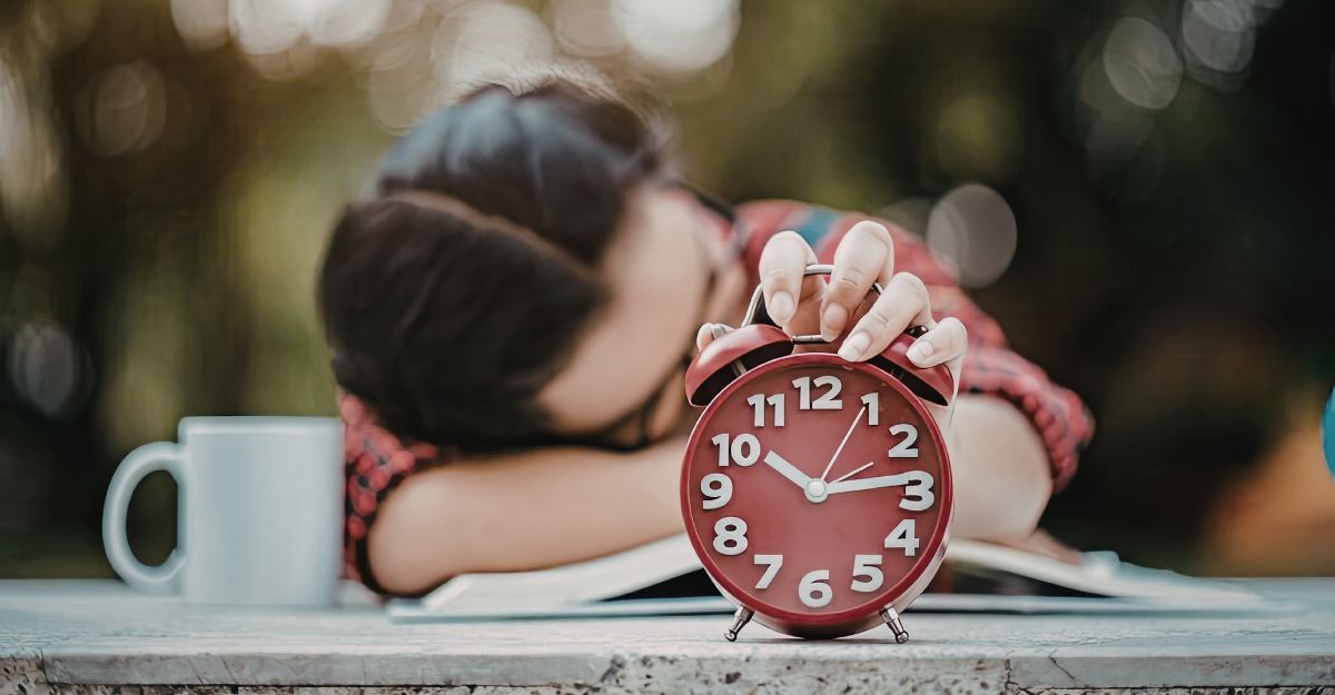 Una persona addormentata su un tavolo accanto a una tazza, con la mano appoggiata su una sveglia rossa
