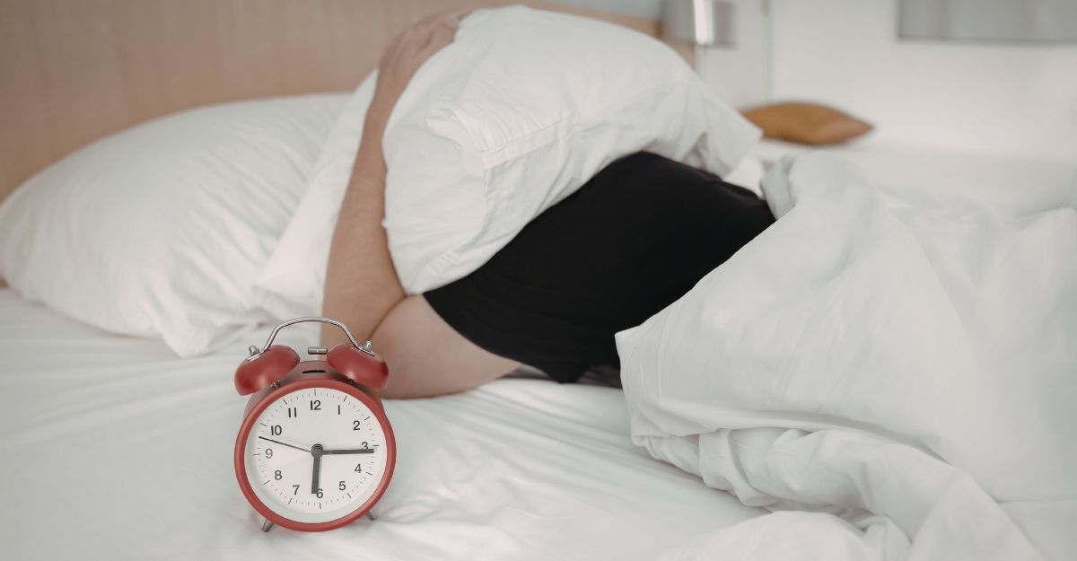 Una persona a letto che si copre la testa con il cuscino accanto a una sveglia rossa