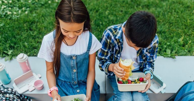 Fratello e sorella minore seduti sull'erba in un parco estivo, con i loro portapranzi al sacco e lo smartwatch al polso