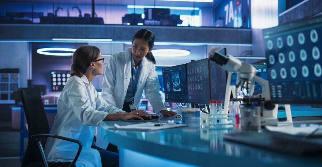 Dottoresse asiatiche e caucasiche parlano in un centro di ricerca medica e utilizzano un computer per analizzare scansioni TC cerebrali. Un chirurgo e un neuroscienziato discutono di soluzioni per il trattamento dei pazienti oncologici.