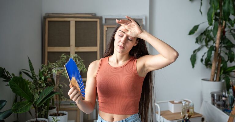 Una giovane donna in un interno luminoso che si rinfresca con un ventaglio a mano e si asciuga il sudore dalla fronte