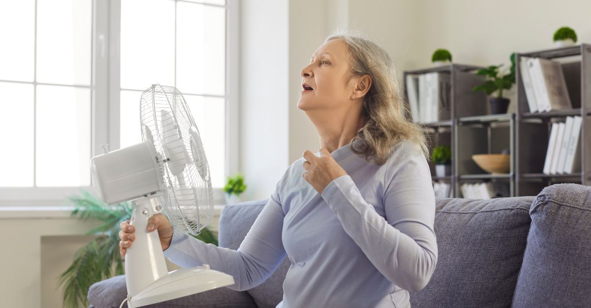 Una donna anziana seduta sul divano che cerca sollievo dal caldo orientando un ventilatore da tavolo verso il proprio viso