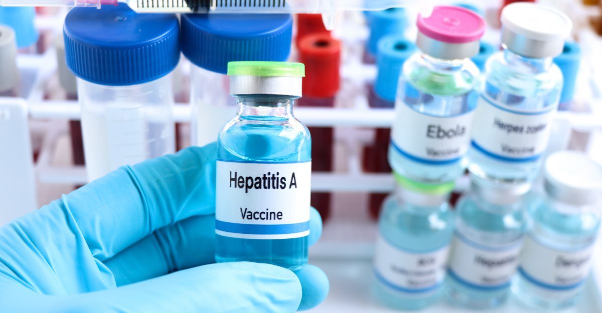 Una mano con guanto azzurro tiene un flacone di vaccino contro l'Epatite A, con altri vaccini sullo sfondo.
