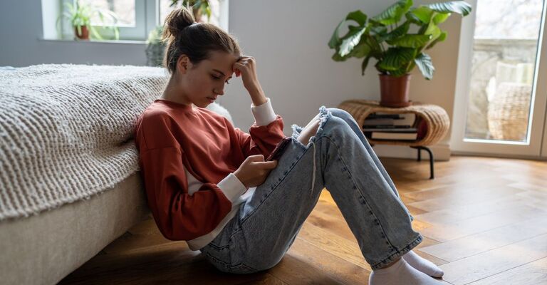 Adolescenti e cyberbullismo. Una ragazza adolescente turbata è seduta sul pavimento vicino al letto e usa lo smartphone a casa, scorrendo i social media. Una bambina passa troppo tempo al telefono. Adolescenti e dipendenza dai dispositivi elettronici.