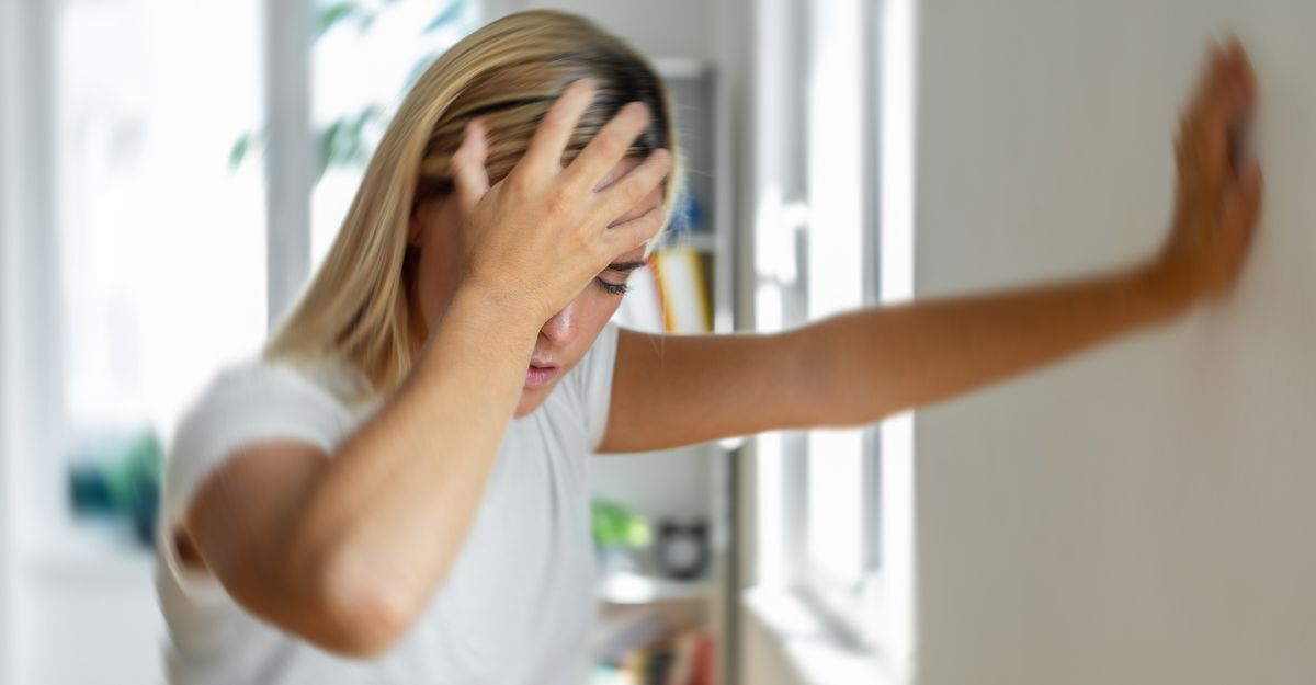 Una giovane donna bionda con un'espressione sofferente si tiene la testa con una mano e si appoggia al muro con l'altra