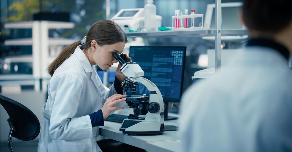 Una scienziata osserva campioni biologici al microscopio in un laboratorio di scienze applicate. Ritratto di un ingegnere di laboratorio caucasico in camice bianco seduto di spalle alla telecamera.