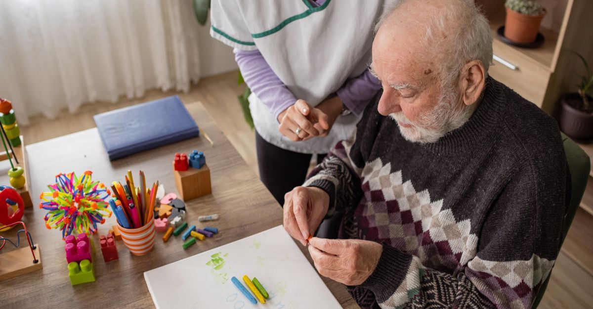 Un uomo anziano impegnato in un'attività di disegno con i pastelli, assistito da un'operatrice sanitaria in una sala luminosa