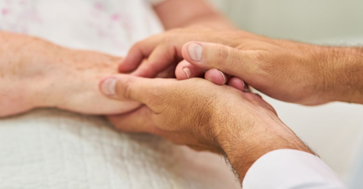 Primo piano di una persona che stringe con cura e calore le mani di un paziente anziano disteso a letto.