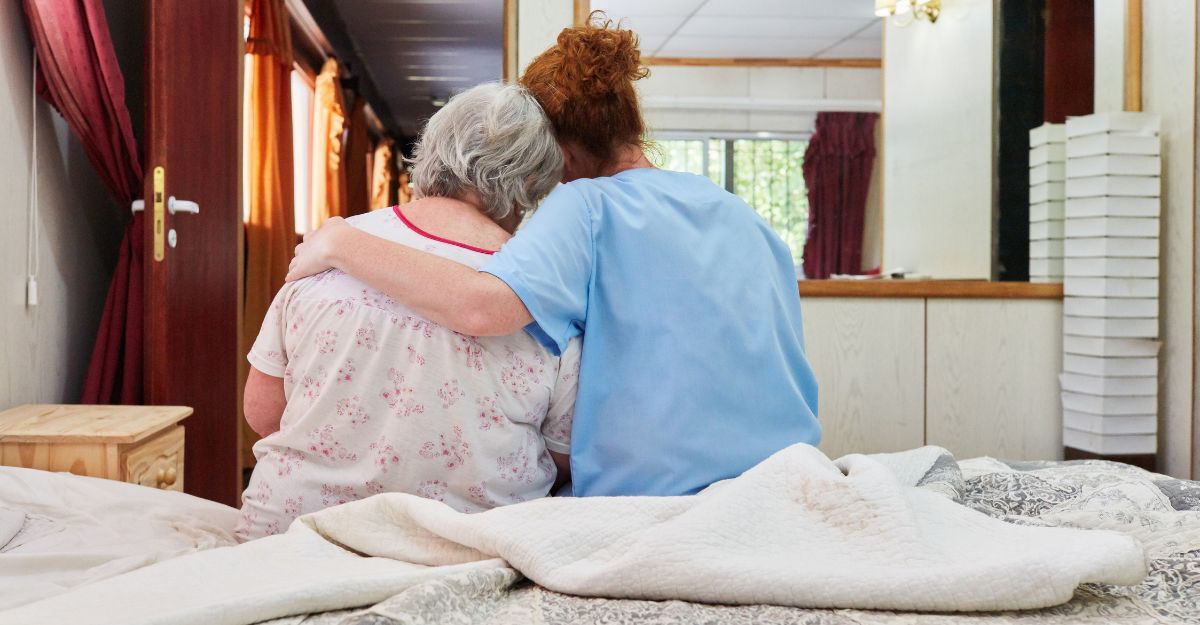 Un'operatrice sanitaria abbraccia affettuosamente una donna anziana seduta sul letto, offrendo conforto in un momento di vicinanza.