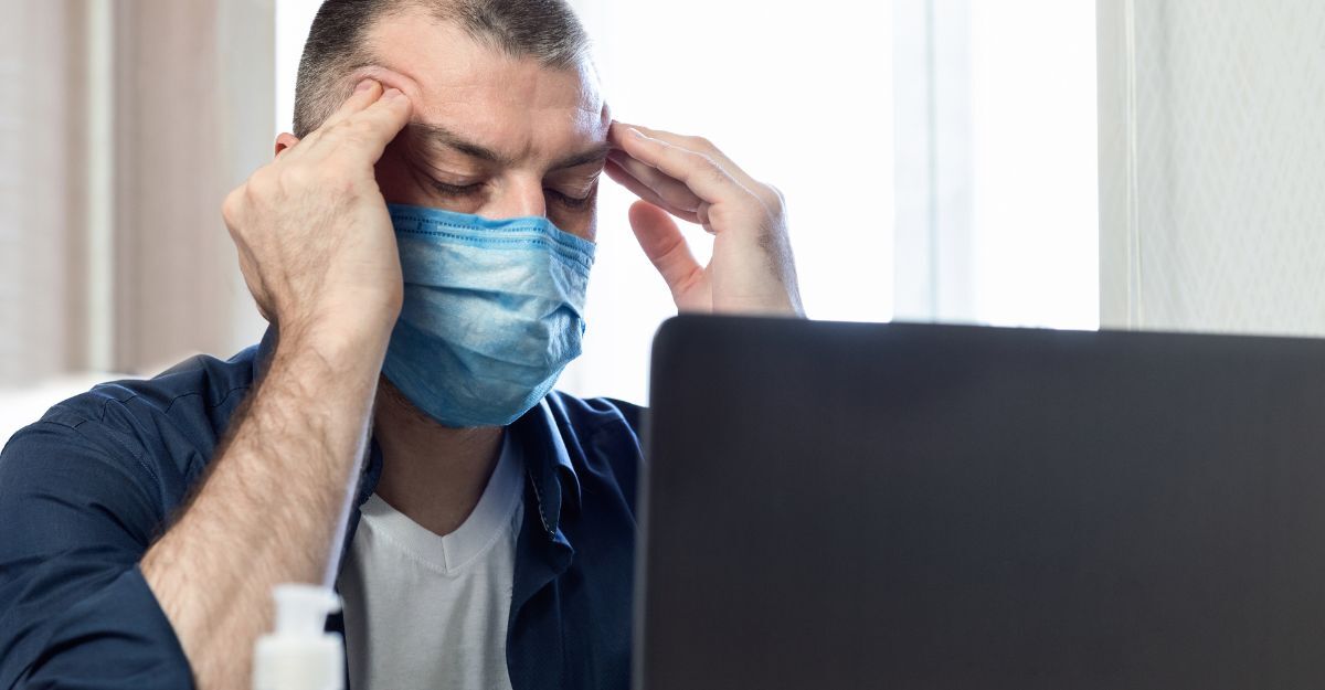 Un uomo con mascherina protettiva seduto davanti a un laptop mentre si tocca le tempie con espressione sofferente