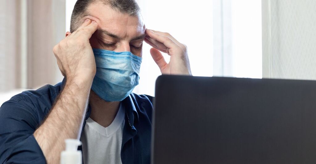 Un uomo con mascherina protettiva seduto davanti a un laptop mentre si tocca le tempie con espressione sofferente