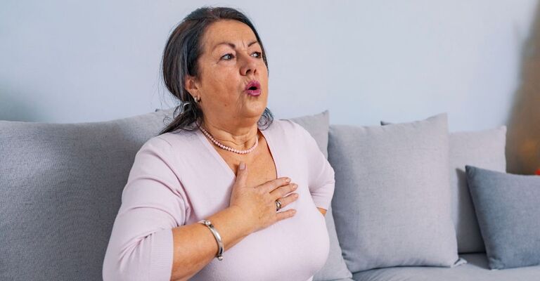 Donna seduta sul divano con espressione sofferente e la mano poggiata sul petto.