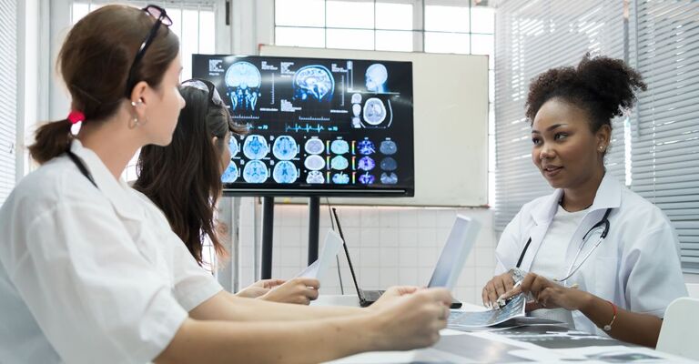 Un'équipe di medici si riunisce nel laboratorio di ricerca sul cervello, con un monitor che mostra immagini di risonanza magnetica e TAC cerebrali. Un gruppo di dottoresse discute di interventi chirurgici e trattamenti, con l'ausilio di radiografie.