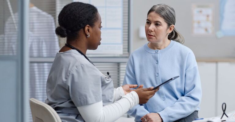 Un'infermiera discute informazioni mediche con una donna caucasica di mezza età in un contesto clinico; l'infermiera tiene in mano un tablet digitale mentre la paziente ascolta attentamente durante la consultazione.