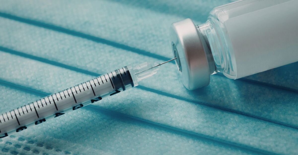 Una siringa che preleva del liquido da una fiala medica, appoggiate su una mascherina chirurgica azzurra