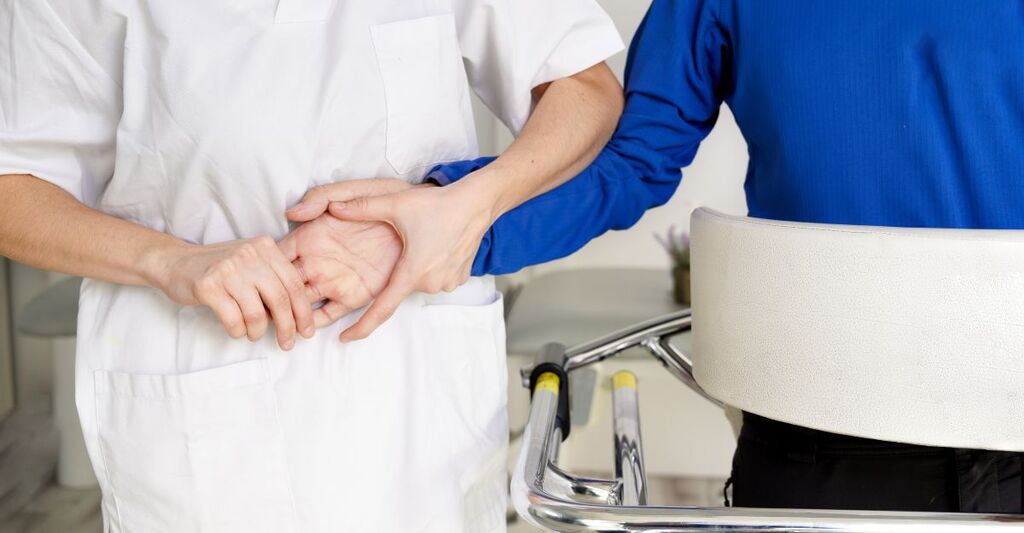 Primo piano di un operatore sanitario che sostiene con cura il braccio e la mano di un paziente vicino a un deambulatore.