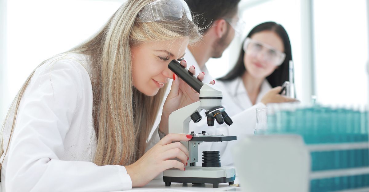 Una ricercatrice in camice bianco osserva un campione al microscopio in un laboratorio, con colleghi sfocati sullo sfondo.