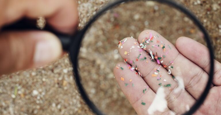 Una mano che regge frammenti di microplastiche osservata attraverso una lente d'ingrandimento su uno sfondo di sabbia