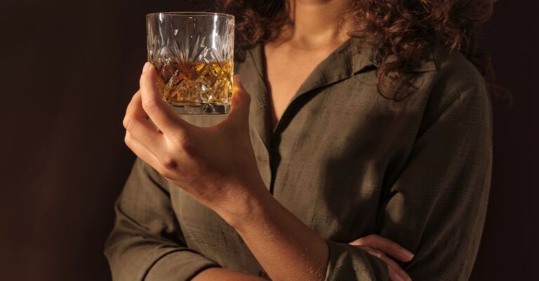 Una persona tiene in mano un bicchiere di cristallo con del whiskey su uno sfondo scuro.