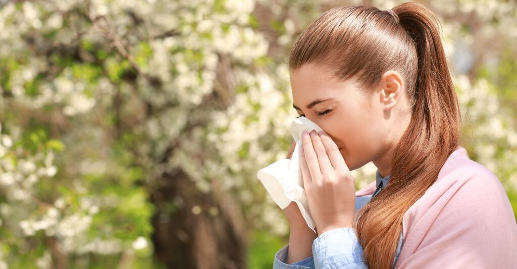 Donna che starnutisce con un fazzoletto per il naso tra gli alberi in fiore in un parco