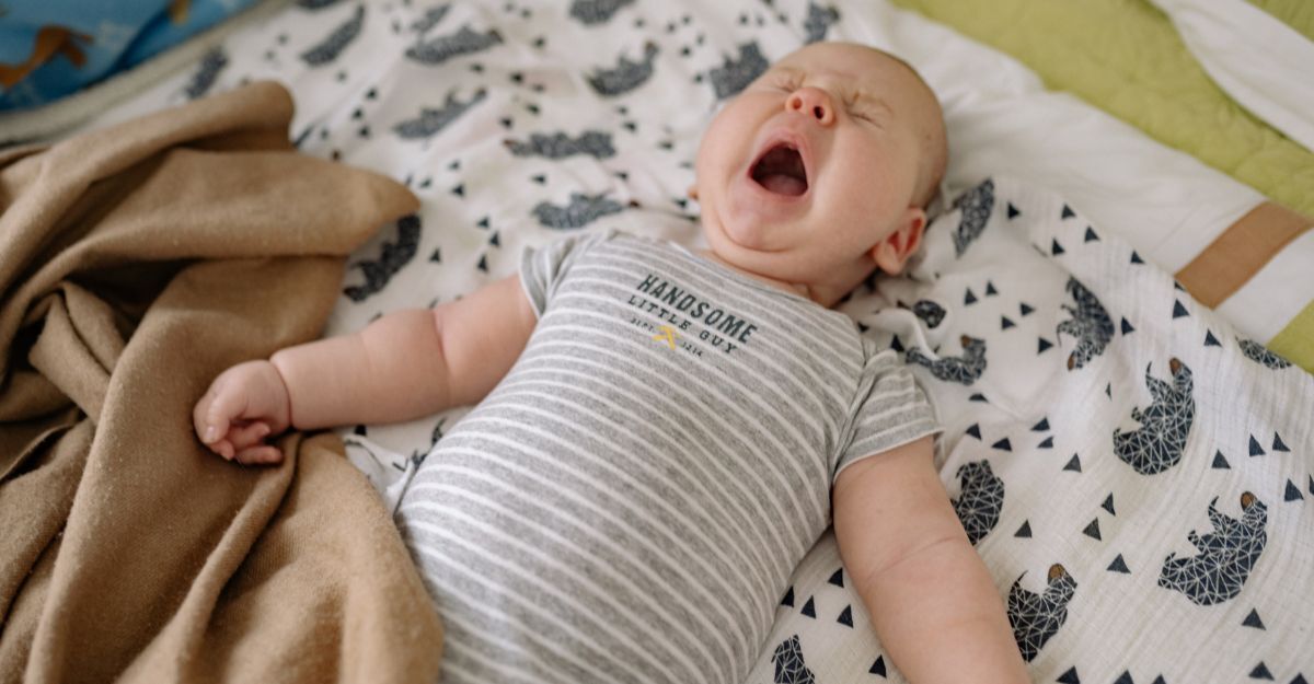 Neonato con tutina a righe che sbadiglia o piange disteso su una copertina a motivi geometrici.