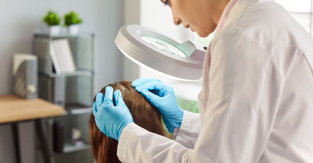 Una specialista in camice bianco e guanti azzurri esamina il cuoio capelluto di una paziente usando una lampada d'ingrandimento professionale