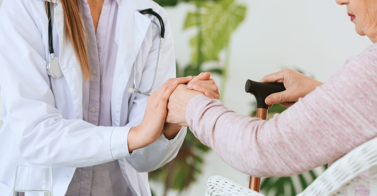 Un medico in camice bianco stringe con empatia le mani di una persona anziana che impugna un bastone da passeggio.