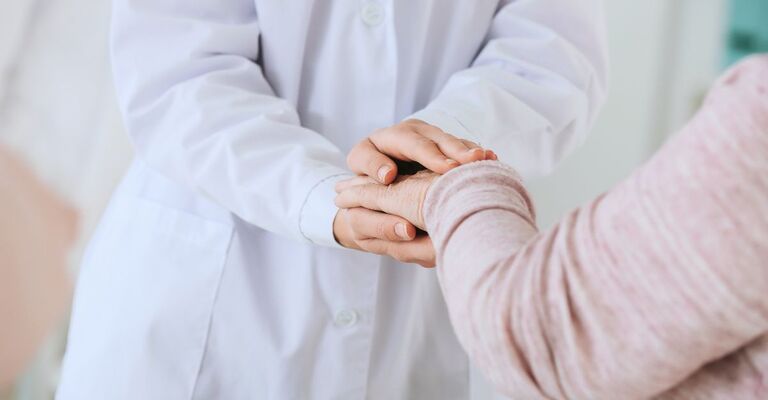 Primo piano di un medico in camice bianco che stringe con cura le mani di un paziente anziano