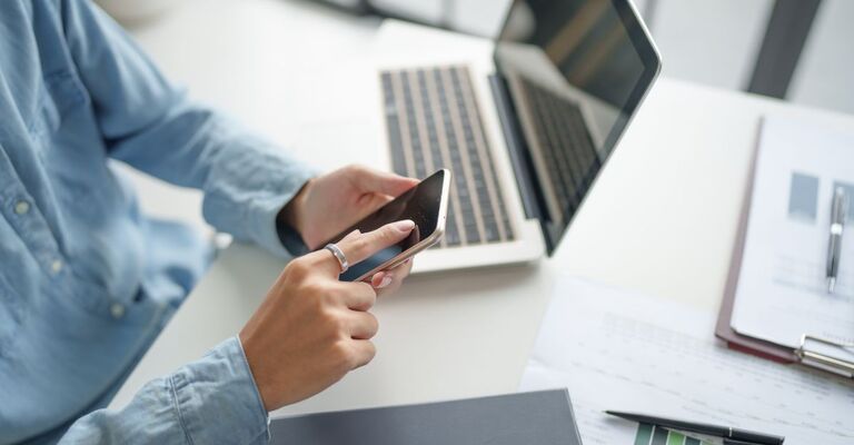 Donna con camicia di jeans azzurra siede a una scrivania, con un computer portatile davanti, mentre utilizza lo smartphone