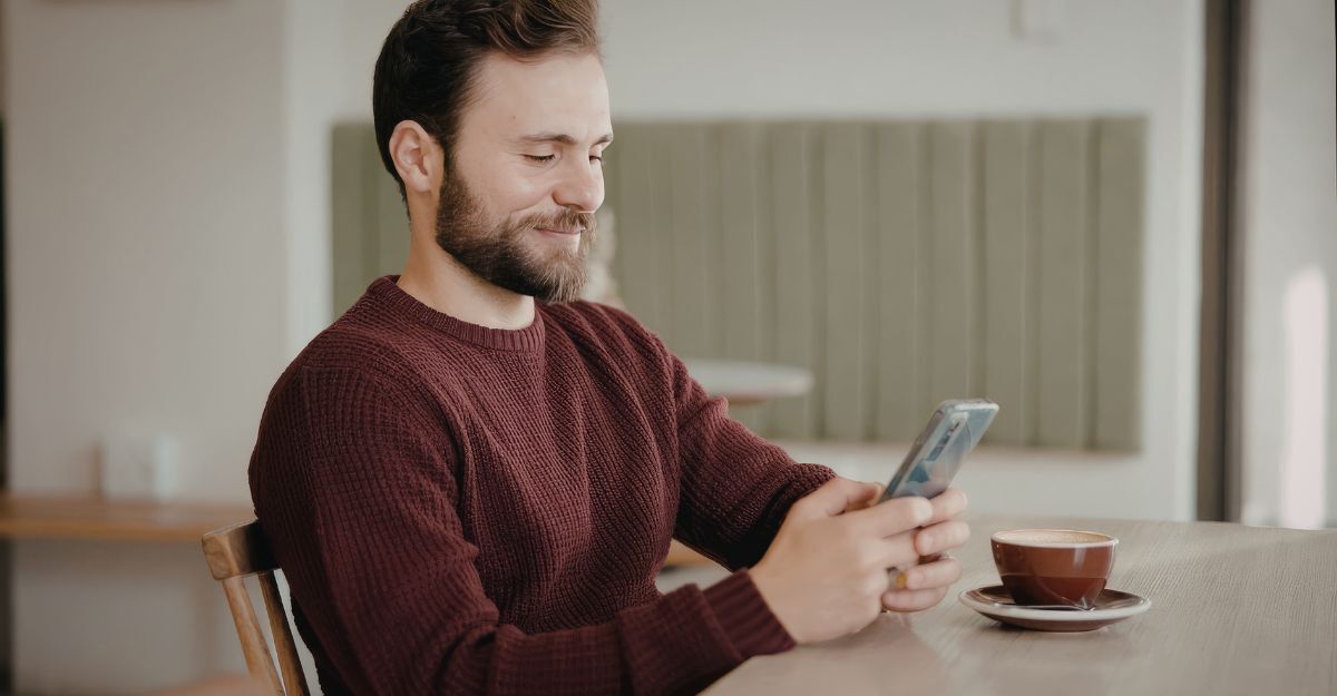 Uomo siede a un tavolo con una tazza di caffè, sorridendo mentre guarda il proprio smartphone