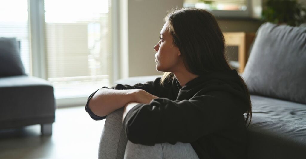 Una donna caucasica triste è seduta sul pavimento e guarda fuori dalla finestra di casa