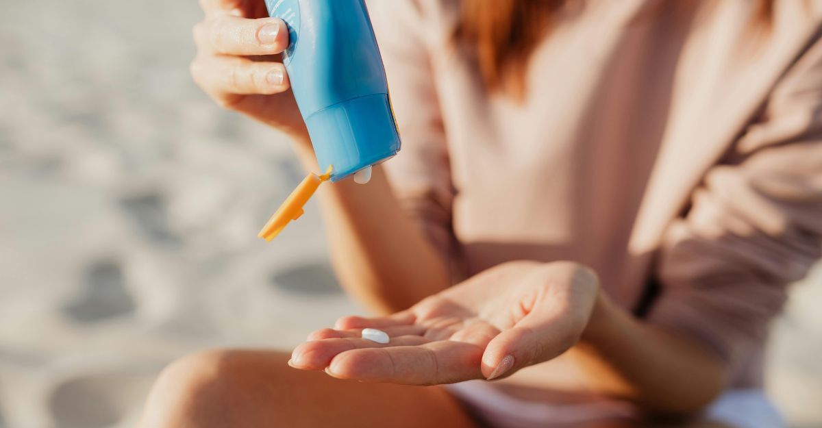 Una donna seduta in spiaggia versa della crema solare dalla bottiglia sul palmo della mano. In primo piano le mani e le gambe