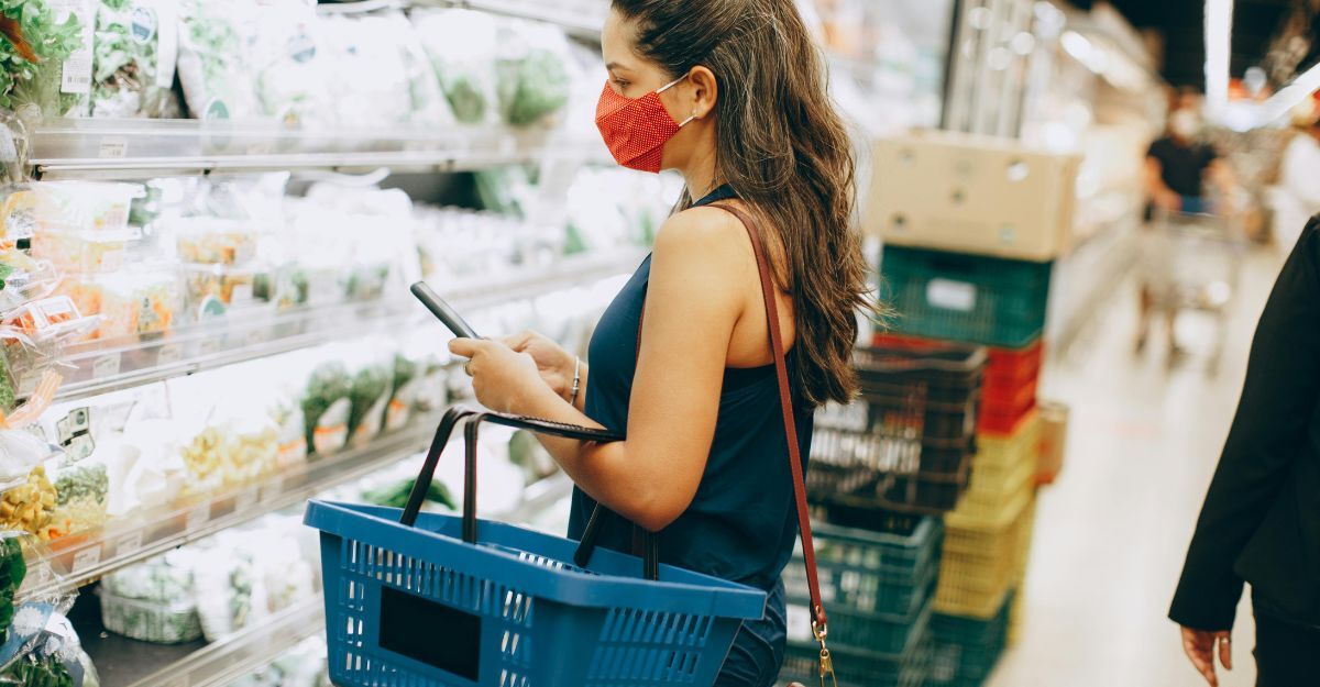 Donna con mascherina rossa che consulta lo smartphone in un supermercato tenendo un cestino della spesa blu