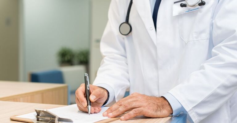 Dettaglio di un medico in camice bianco e stetoscopio mentre scrive con una penna su un modulo sopra una cartella clinica a molla