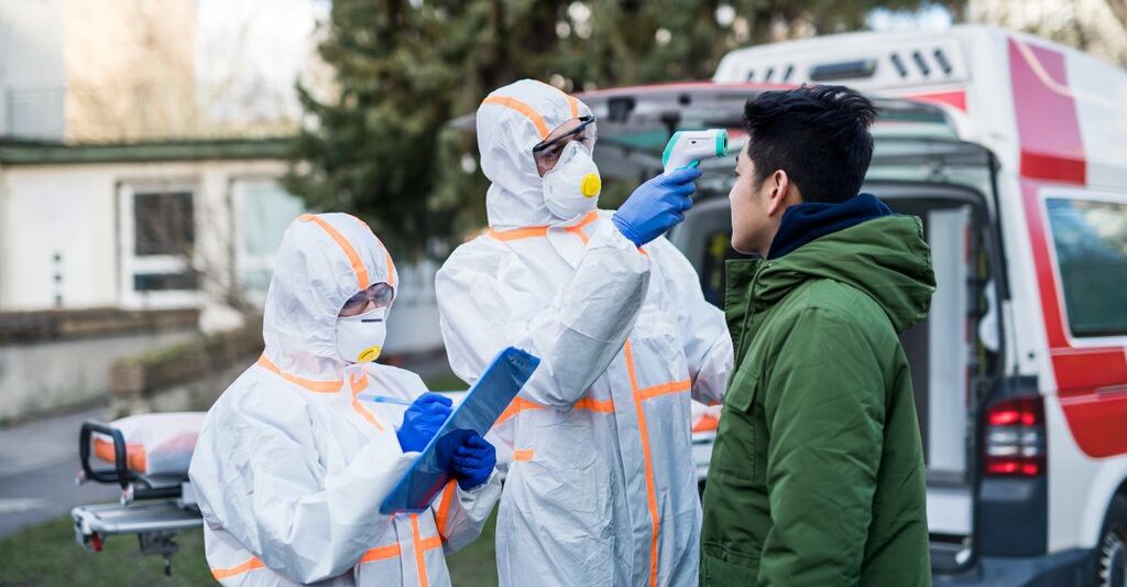 Personale sanitario in tuta protettiva e mascherina mentre misura la febbre a un uomo con un termometro a infrarossi davanti a un'ambulanza