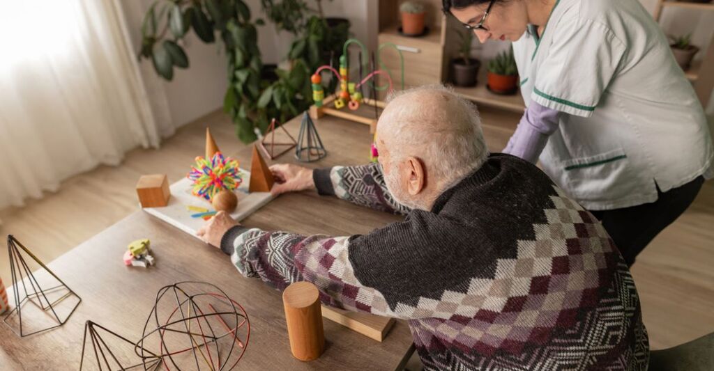 Un anziano seduto a un tavolo mentre maneggia forme geometriche in legno, assistito da un'operatrice sanitaria di fianco a lui.