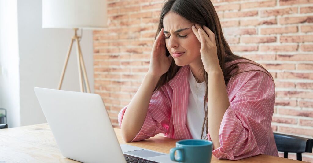 Donna seduta davanti al laptop con le mani sulle tempie ed espressione di stress o mal di testa