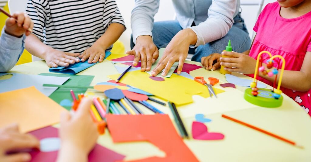 Bambini e un'adulta seduti a un tavolo mentre fanno lavoretti creativi con carta colorata, forbici e matite