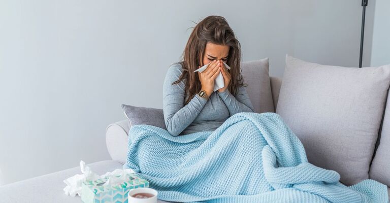 Donna sul divano coperta da una coperta azzurra che si soffia il naso, con una scatola di fazzoletti accanto.