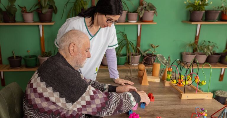Un'operatrice socio-sanitaria assiste un uomo anziano seduto a un tavolo mentre svolge attività di stimolazione cognitiva con blocchi e giochi di legno.   