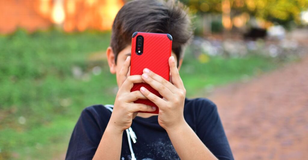 Ragazzo all'aperto che copre il proprio viso tenendo lo smartphone rosso in verticale davanti a sé