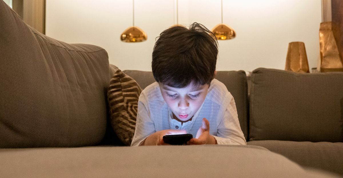 Bambino disteso sul divano al buio che guarda attentamente lo schermo del suo smartphone illuminato