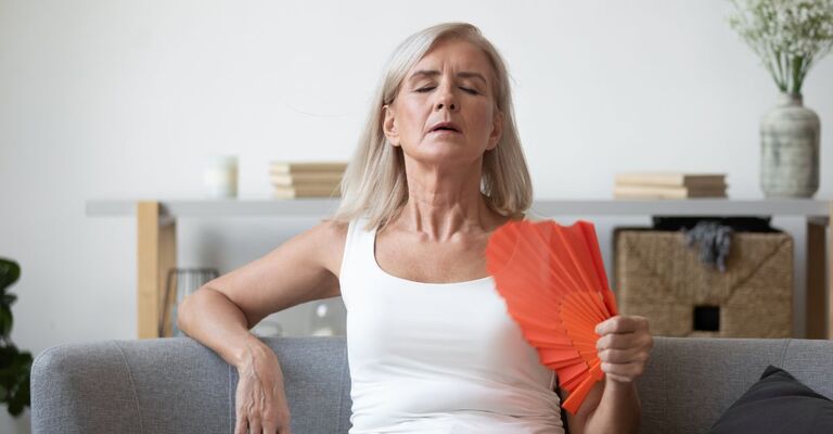 Donna seduta sul divano che cerca di rinfrescarsi con un ventaglio arancione, soffrendo manifestamente la vampata di calore legata alla menopausa
