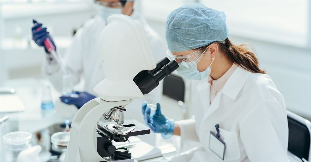 Una ricercatrice in camice bianco, guanti protettivi, mascherina e occhiali da protezione mentre osserva un campione al microscopio all'interno di un laboratorio di analisi