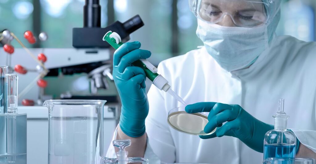 Ricercatore in tenuta protettiva completa con mascherina e occhiali che utilizza una micropipetta per applicare un campione in una capsula di Petri