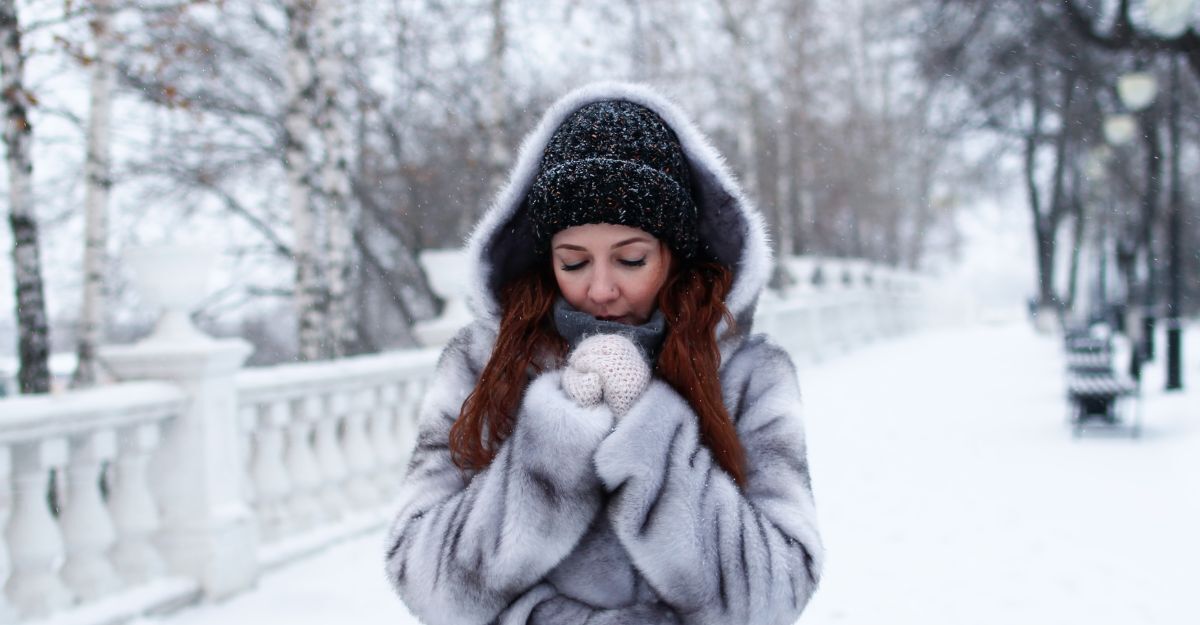 Una donna ben coperta con cappello, guanti e cappotto con cappuccio che si scalda le mani in un paesaggio innevato