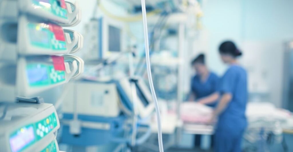 Vista ravvicinata di apparecchiature mediche e pompe d'infusione in una stanza d'ospedale, con due operatori sanitari sfocati sullo sfondo.